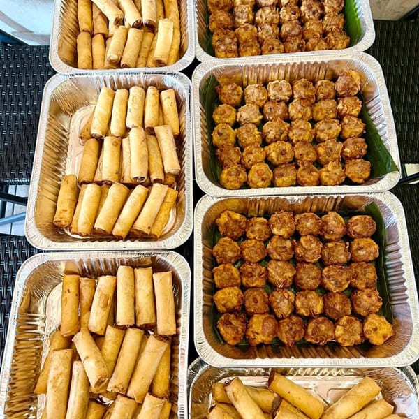 Multiple foil trays filled with golden-brown spring rolls and fried dumplings on banana leaves.