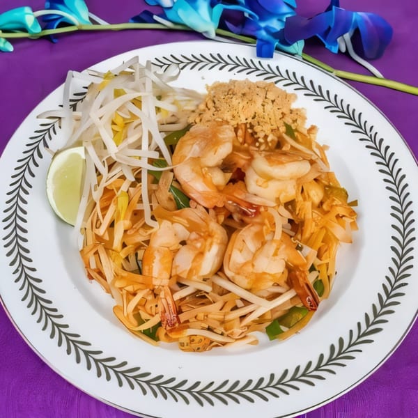 Shrimp Pad Thai with bean sprouts, lime, and crushed peanuts on a white plate with a purple background.