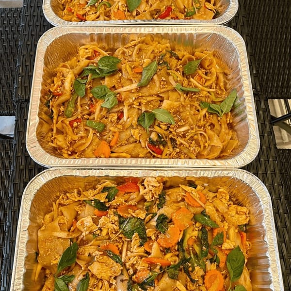 Three aluminum trays filled with Pad See Ew noodles, chicken, vegetables, and basil leaves.