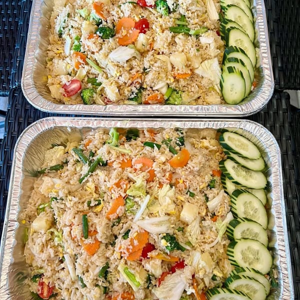 Two aluminum trays filled with vegetable fried rice and garnished with cucumber slices.