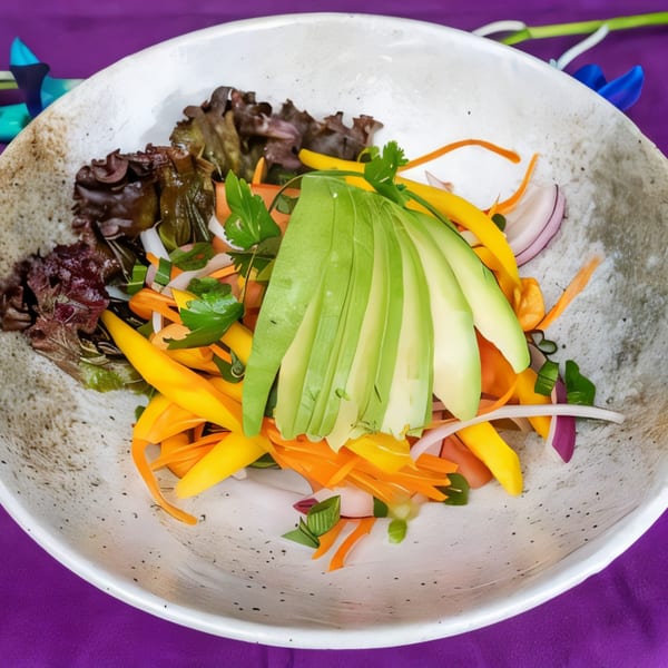 Colorful salad with sliced avocado, mango, carrots, red onion, and greens in a white bowl.